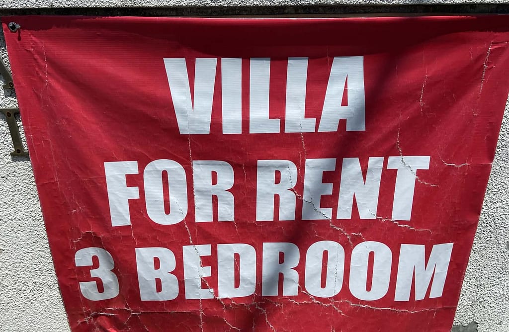 Červená oprýskaná plachta s nápisem Villa for rent 3 bedroom zavěšená na zdi na Bali.