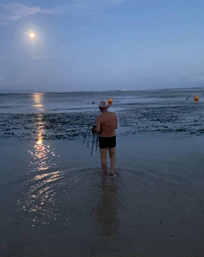 Muž stojící v mělké vodě během večerního odlivu za úplňku na pobřeží v Sanuru na Bali.